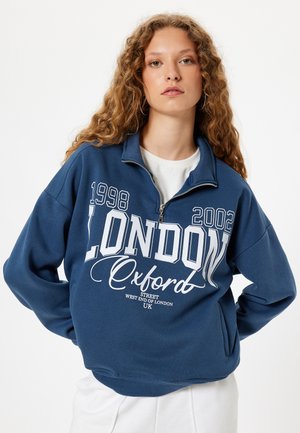 Sudadera de color azul oscuro con un alto cuello, cierre de cremallera, texto gráfico blanco que presenta "LONDRES" y "Oxford", y puños y dobladillo de canalé.
