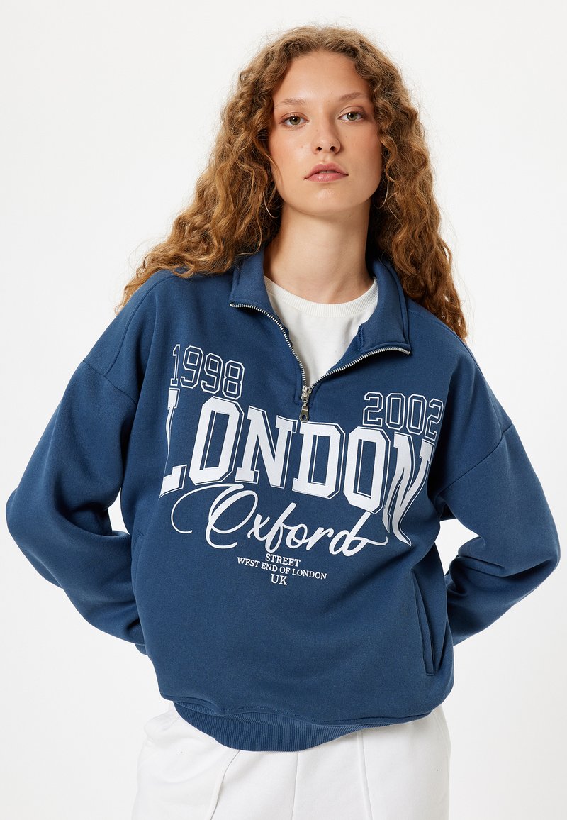 Sudadera de color azul oscuro con un alto cuello, cierre de cremallera, texto gráfico blanco que presenta "LONDRES" y "Oxford", y puños y dobladillo de canalé.