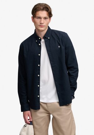 Joven con una camisa azul marino de botones sobre una camiseta blanca y pantalones beige, sosteniendo una gorra beige en su mano izquierda.