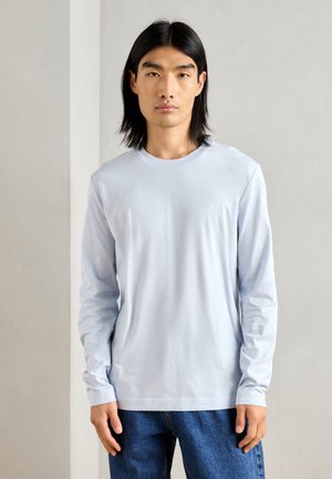 T-shirt à manches longues bleu clair en tissu doux avec un col ras du cou et un détail de logo subtil sur la poitrine. Associé à un denim.