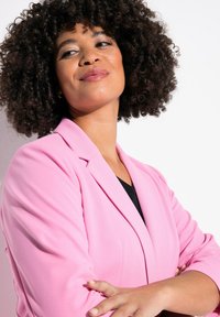 Blazer rosa con una textura suave, corte entallado y solapas anchas. La modelo lleva un top negro debajo, mostrando un aspecto elegante y profesional.