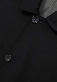 Mantou negru cu un guler structurat, două butoane mari și o textură de țesătură netedă, evidențiind un design minimalist.
