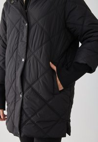 Schwarze gesteppte Oberbekleidung mit Diamantmustern, Druckknöpfen und gerippten Ärmeln. Verfügt über Seitentaschen und ein hochgeschlossenes Design.