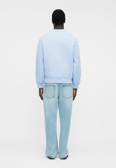 Άτομο που στέκεται με την πλάτη γυρισμένη, φοράει ανοιχτομπλε φούτερ και φαρδιά ανοιχτομπλε τζιν, σε απλό φόντο.