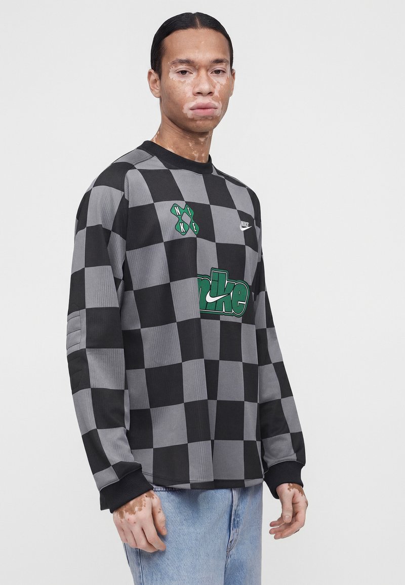 Junge Person mit Vitiligo trägt schwarzen und grauen karierten Nike-Sweatshirt und hellblaue Jeans, steht vor weißem Hintergrund.