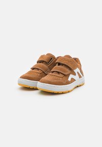 Chaussures en daim marron avec des accents blancs, dotées d'une surface texturée, de deux bandes Velcro et d'une semelle extérieure blanche avec un fond en caoutchouc jaune.