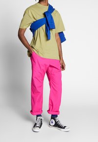 Person som bär ljusrosa byxor, beige t-shirt, en blå sweatshirt knuten runt axlarna och svarta högtopps Converse-skor.