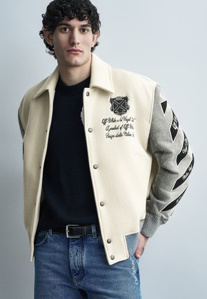 Joven con cabello rizado que lleva una chaqueta universitaria crema y gris con texto y gráficos en negro, combinada con un suéter negro y jeans azules.