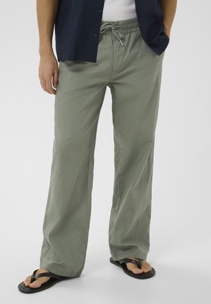 Dolna część ciała osoby noszącej luźne, jasnozielone spodnie ze sznurkiem, czarne klapki, biały top oraz granatową koszulę, z rękami w kieszeniach.