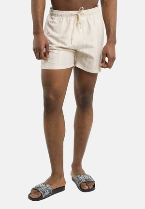 Mand iført beige badeshorts med snøre og sorte sandaler med mønstrede remme, stående med benene let adskilte.