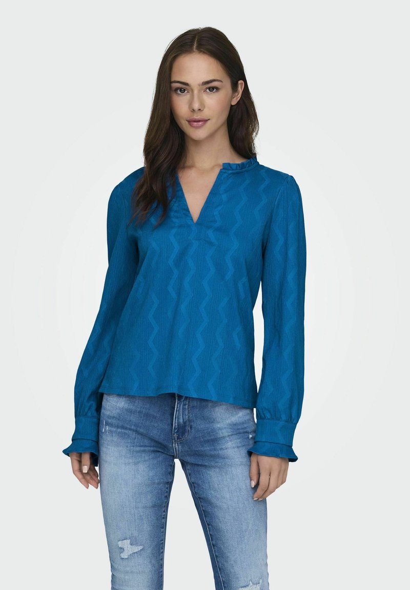 Blouse bleue texturée avec un décolleté en V, un col à volants et des manches longues, associée à un jean déchiré. Présente un motif en zigzag.