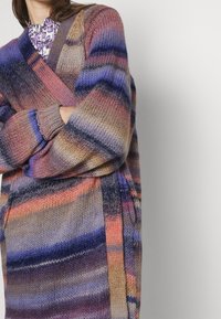 Sweter z dzianiny w gradientowych odcieniach fioletu, różu i niebieskiego. Posiada fason kopertowy oraz szerokie rękawy z prążkowanymi mankietami.