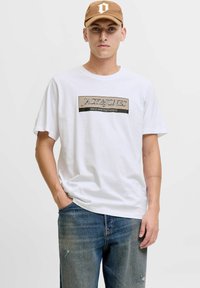 Camiseta de algodón blanca con un logo impreso en negro y beige, combinada con unos jeans de mezclilla azul descoloridos y una gorra marrón.