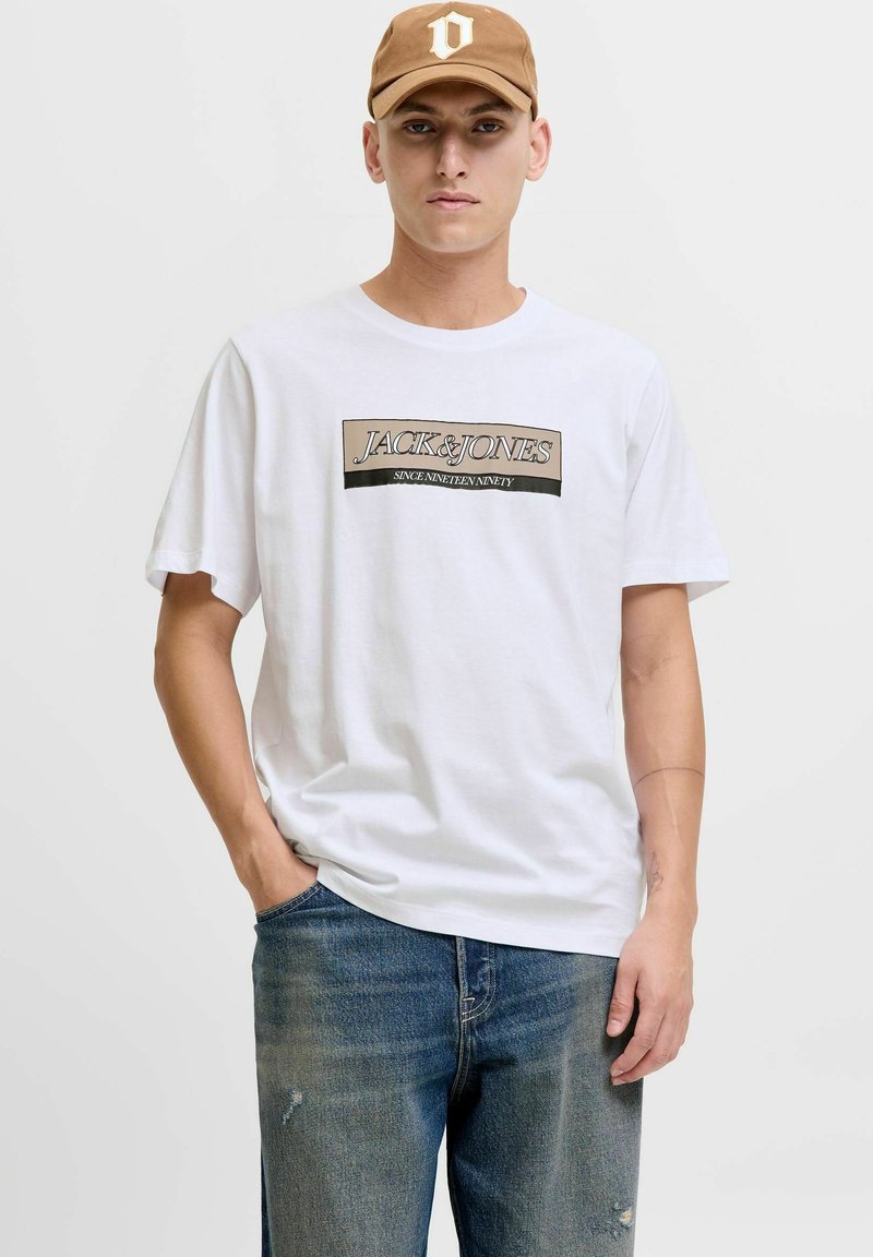 Camiseta de algodón blanca con un logo impreso en negro y beige, combinada con unos jeans de mezclilla azul descoloridos y una gorra marrón.