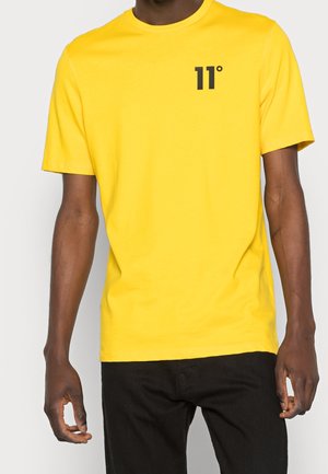 Geel katoenen T-shirt met een zwart "11°" grafisch ontwerp op de linkerborst, korte mouwen en een relaxte pasvorm. Gepaarde met zwarte broek.