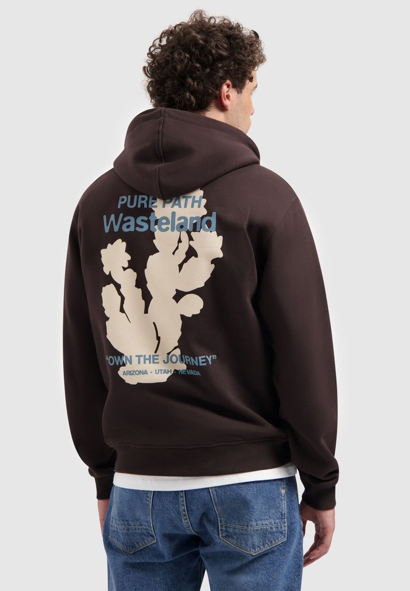 Man in een donkerbruine hoodie met een cactusafbeelding en de tekst "Pure Path Wasteland Own the Journey Arizona Utah Nevada" op de achterkant.