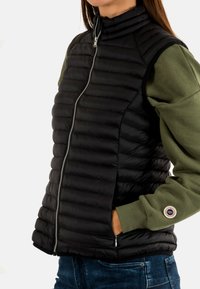 Chaleco acolchado negro ligero con un cuello alto, cierre de cremallera y un diseño acolchado. Llevado sobre una camiseta de manga larga verde con un parche de logo.