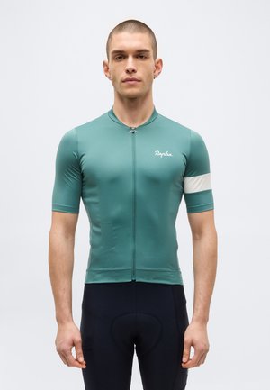 Moški, ki nosi zeleno kratkoročno kolesarsko majico z belo črto na roki in črne oblazinjene kolesarske hlače, stoji ob enobarvnem ozadju.