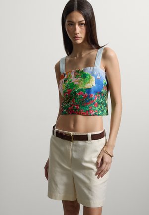 Junge Frau mit langen dunklen Haaren, die ein Crop-Top mit floralem Landschaftsprint und cremefarbene, hoch taillierte Shorts mit braunem Gürtel trägt.