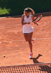 Female tennis player in white dress hitting a ball on a red clay court during a sunny match.
