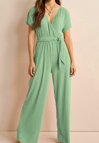 Combinaison plissée verte avec un design enroulé, des manches courtes, une taille nouée et des jambes larges, offrant une texture lisse et un style minimaliste.