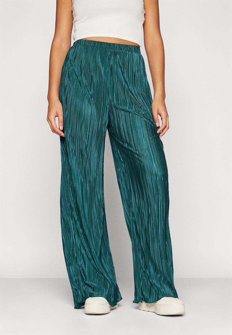 Femme portant un pantalon large plissé bleu sarcelle, un top court blanc et des baskets blanches à plateforme, debout devant un fond uni.