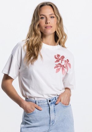 Blonde vrouw met golvend haar, dragend een wit T-shirt met roze bloemenborduursel en lichtblauwe high-waisted jeans, handen in de zakken.