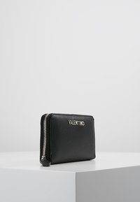 Portefeuille en cuir noir avec une surface texturée, fermeture à zip et logo doré. Forme rectangulaire compacte, affichant un profil mince.