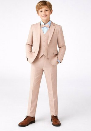 Garçon souriant les mains dans les poches, portant un costume trois pièces beige, une chemise bleu clair, un nœud papillon beige et des chaussures marron habillées, debout sur fond blanc.