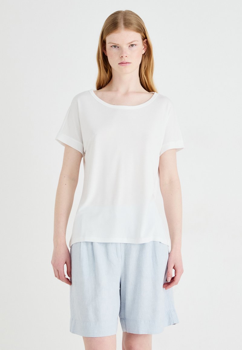 Haut blanc à manches courtes avec un col rond et des poignets retroussés, fabriqué en tissu lisse. Associé à un short en lin bleu clair, ample.