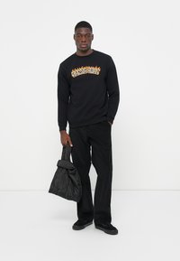 Zwarte sweatshirt met oranje vlamrand rondom de tekst "SANTACRUZ", gecombineerd met zwarte wijde broek en een gewatteerde zwarte tas.