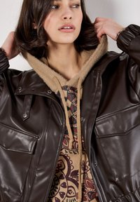 Chaqueta de cuero sintético marrón con botones a presión, dos bolsillos en el pecho y una sudadera con capucha forrada de felpa color beige con un patrón de paisley debajo.