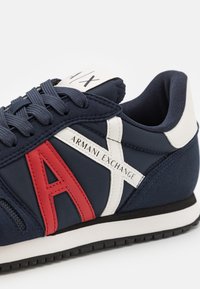 Laivastonsininen lenkkikenkä valkoisilla ja punaisilla nahkaosilla, laivastonsinisillä nauhoilla, "Armani Exchange" -merkinnällä sivussa ja mustalla kumipohjalla.