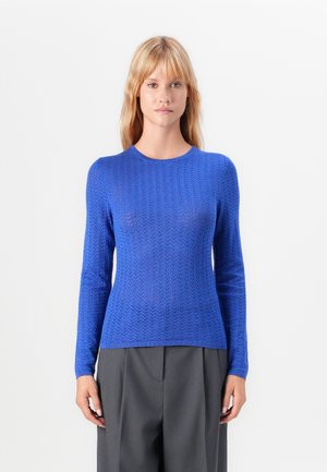 Femme portant un pull ajusté à manches longues bleu texturé et un pantalon gris taille haute, se tenant devant un fond blanc uni.