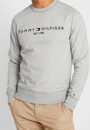 Hellgraues Sweatshirt mit dem Schriftzug "Tommy Hilfiger Est. 1985" und Logo auf der Brust, getragen mit beigen Hosen, Hände in den Taschen.