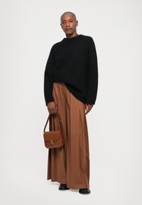 Henkilö seisoo päällään musta neule, leveät ruskeat laskoshousut, mustat kengät, pitelee ruskeaa käsilaukkua valkoista taustaa vasten.