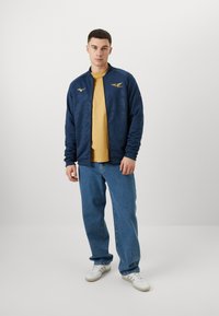 Veste de bombardier marine avec un motif texturé, logo doré et fermeture éclair, portée sur un t-shirt jaune, associée à un jean bleu et des baskets blanches.