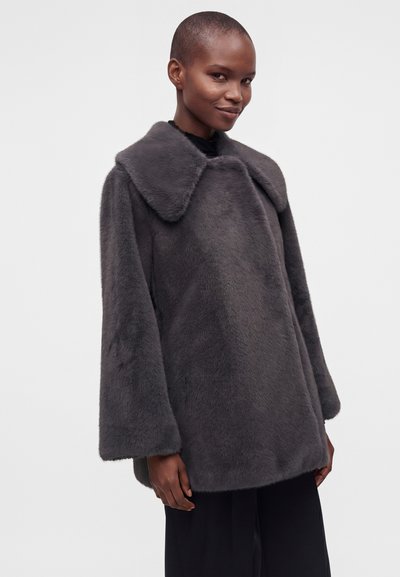 Femme portant un manteau oversize en fausse fourrure gris foncé avec un large col et des manches amples sur un fond clair uni.