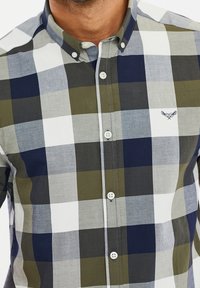 Camicia con bottoni in un motivo a quadri che presenta tonalità di oliva, blu navy, grigio e bianco. Tessuto morbido con colletto arrotondato e bottoni bianchi.