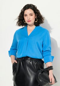 Blusa azzurra a maniche lunghe con scollo a V e dettagli plissettati, abbinata a una gonna di pelle nera e a una borsa nera.