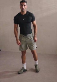 Sort tætsiddende kortærmet skjorte, lysegrå shorts med snøre, hvide sokker og grønne sportssko; neutral baggrund.