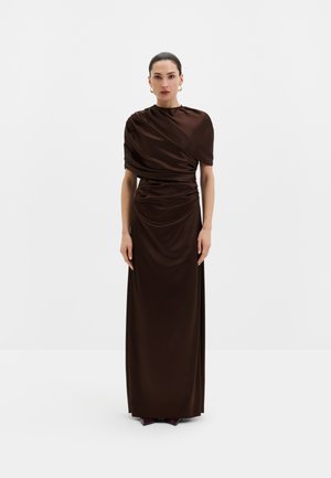 Femme aux cheveux foncés coiffés en chignon lisse porte une robe longue brillante marron foncé avec des détails drapés et des boucles d’oreilles créoles dorées sur fond blanc.
