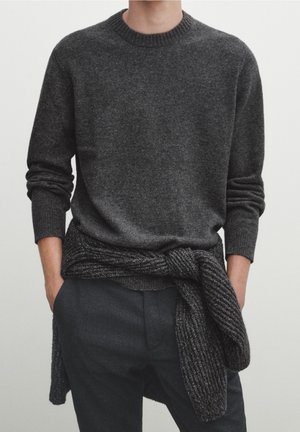 Man wears dark gray crewneck sweater with matching knit cardigan tied around waist and dark pants, hands in pockets.