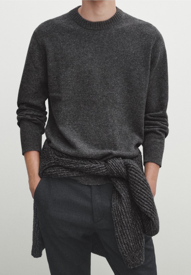 Homme porte un pull gris foncé à col rond avec un cardigan en maille assorti noué autour de la taille et un pantalon foncé, les mains dans les poches.