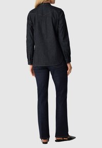 Donkerdenim overhemd met lange mouwen, yoke-detail en contraststiksel, gecombineerd met donkere rechte jeans en platte zwarte sandalen.