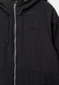 Veste à capuche noire avec une fermeture éclair argentée à l'avant, des cordons de serrage, et un petit logo brodé "SWITZ" sur la poitrine.