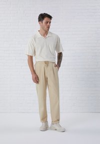 Pull beige à manches courtes avec poche, associé à un pantalon beige à cordon de serrage. Le modèle porte des baskets claires. Tons lisses et neutres.