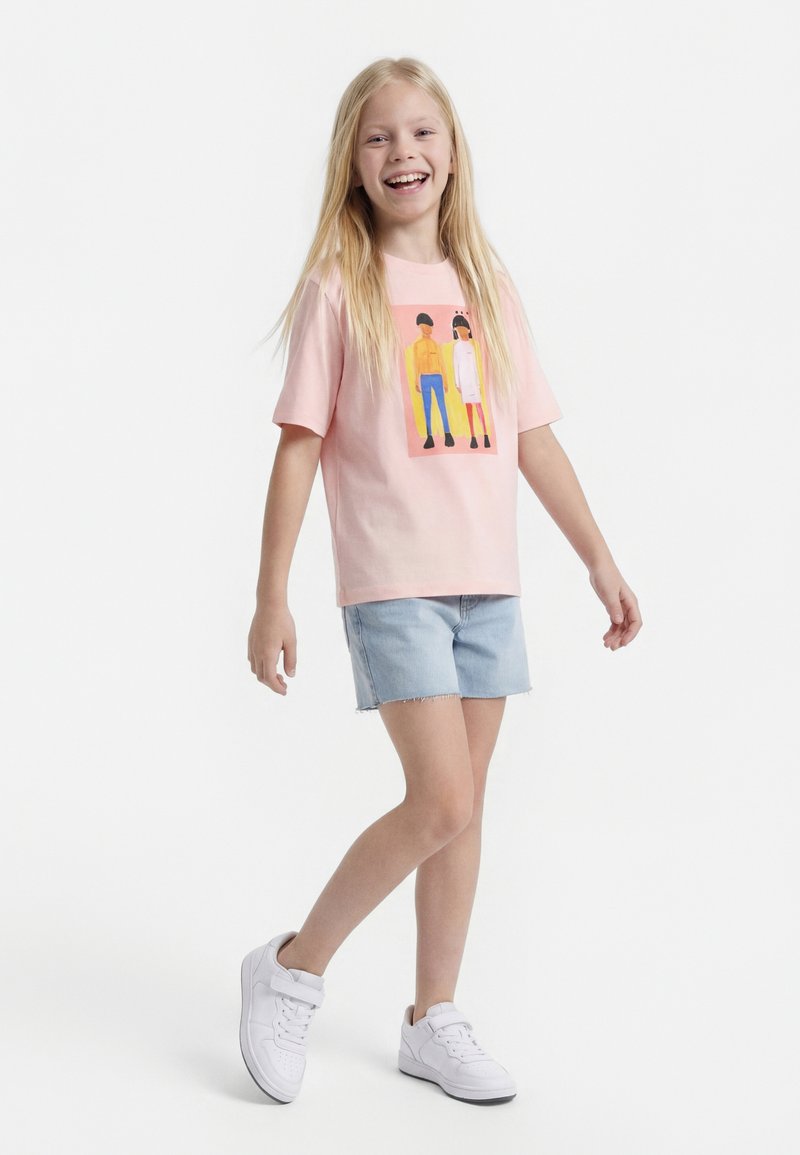 Jeune fille souriante aux longs cheveux blonds, portant un t-shirt rose avec un motif graphique, un short en denim clair et des baskets blanches, debout sur un pied.