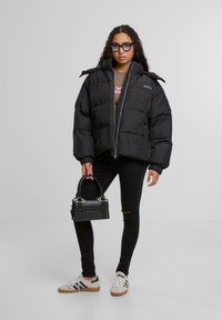 Donna con lunghi capelli ricci che indossa un piumino nero, jeans skinny strappati scuri, sneakers bianche e occhiali, mentre tiene in mano una piccola borsa nera.