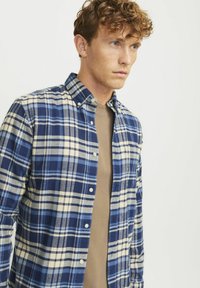 Geruite shirt in marineblauw, crème en lichtblauw, met een button-down kraag en een zachte, textuurstof; gelaagd over een beige shirt.